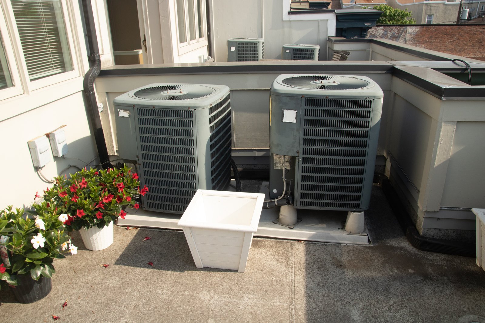 Two outdoor HVAC units installed on a terrace with potted plants nearby, connected to electrical conduits and refrigerant lines.