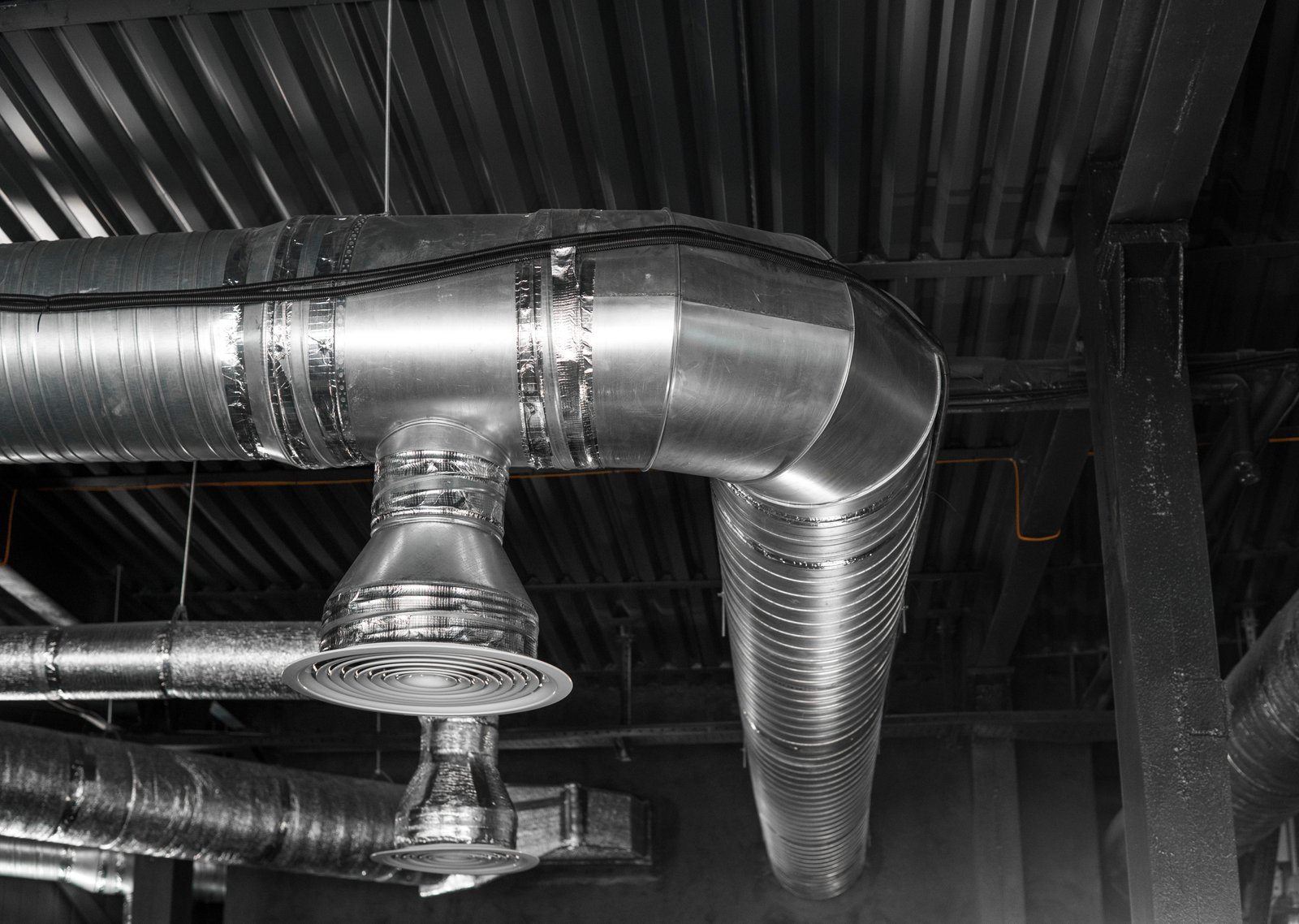 A modern metallic HVAC duct system with large air vents and insulated pipes, installed in an industrial or commercial building.